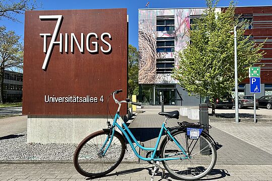 Hellblaues Fahrrad auf dem Parkplatz des Hotels vor dem Eingangsschild und dem Apartmentgebäude im Hintergrund